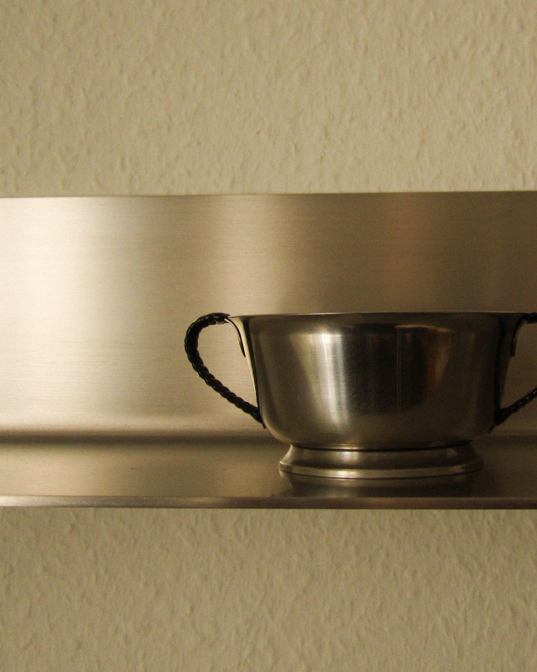 Extreme close-up of a single Skagen shelf holding one vintage-style steel bowl with braided handles. Warm directional light catches both the brushed backplate and the polished bowl surface, creating a tonal dialogue between the two steel finishes. No wall fixings visible. Demonstrates the shelf's depth and load capacity, and how the brushed steel surface complements other metal objects. Strong detail image for articles about styling with steel, material pairings, or the tactile quality of the Skagen shelf surface.