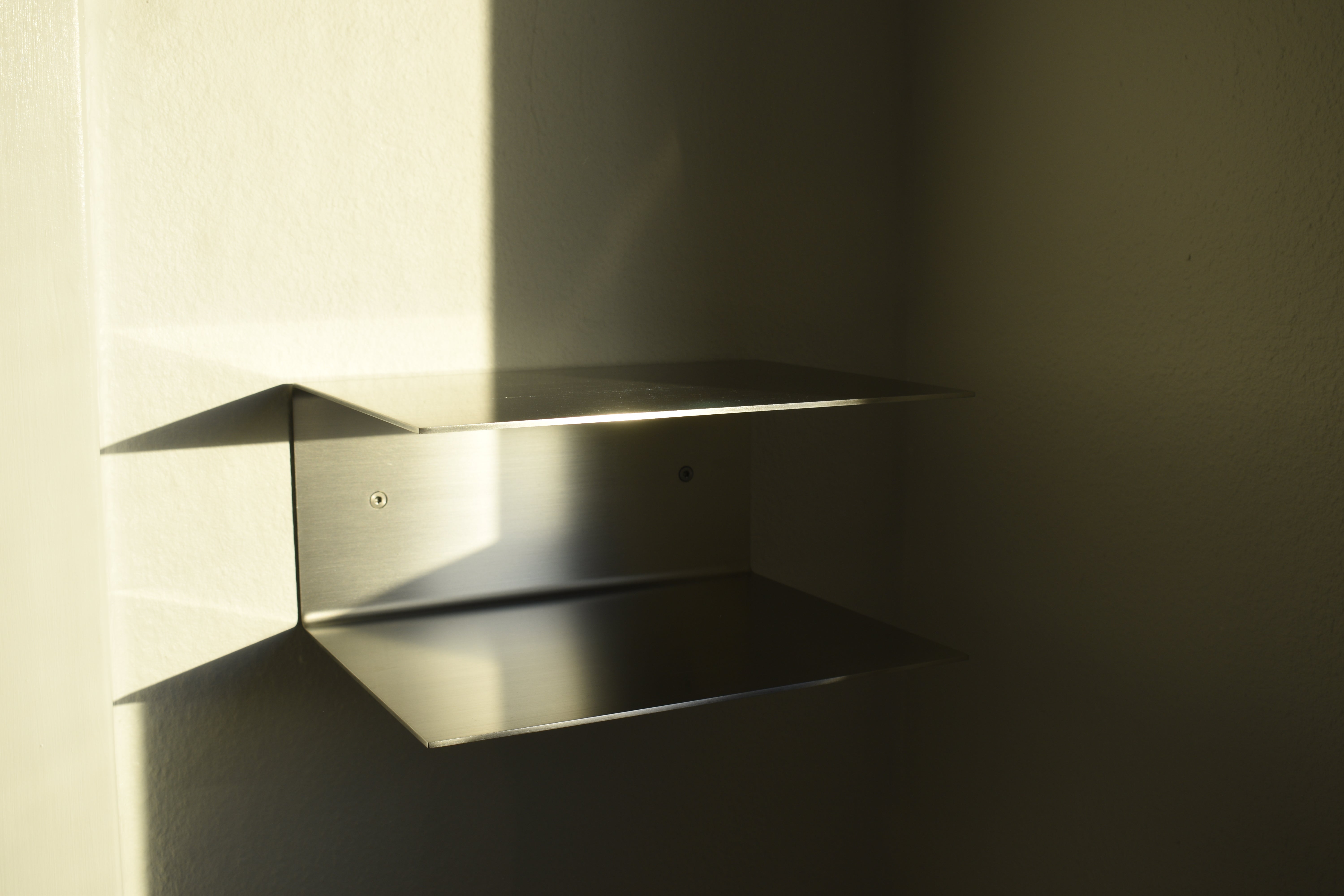Unstyled product shot of the Lund bedside table mounted in a corner where two walls meet. Shot in dramatic directional natural light that highlights the brushed steel surface, the clean folded form and the two visible mounting screws. No objects on the surface. Demonstrates the product's geometric precision and how the piece looks as a pure object, independent of styling.
