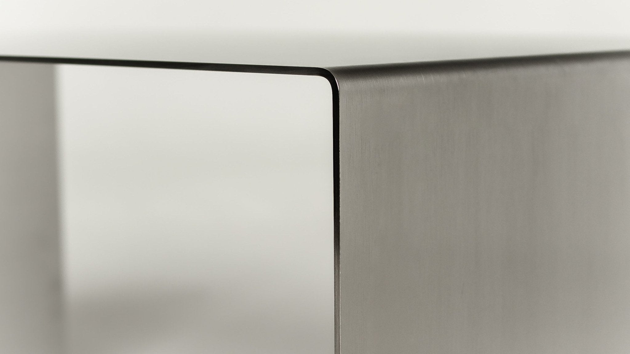 Extreme macro close-up of the Copenhagen coffee table corner, showing the precise folded edge where the top surface meets the side panel. Brushed steel texture clearly visible on the right face, contrasting with the softer reflection on the front panel. The tight radius of the fold and the clean seam line demonstrate the precision of the hand-finishing process. No styling, no context. Best used for articles about craftsmanship, material quality, production precision, or the physical properties of brushed stainless steel.